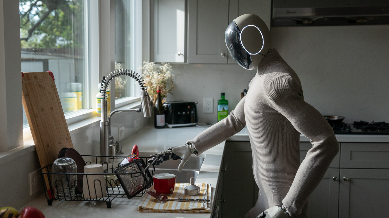 1X NEO Gamma at a dishwasher task demo — home chores sit in analysts' post-2030 wave, while today's money still flows to factories and logistics first.